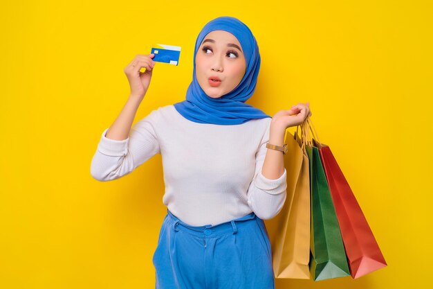 Allegra giovane donna musulmana asiatica che tiene carta di credito e borse della spesa guardando la fotocamera isolata su sfondo giallo