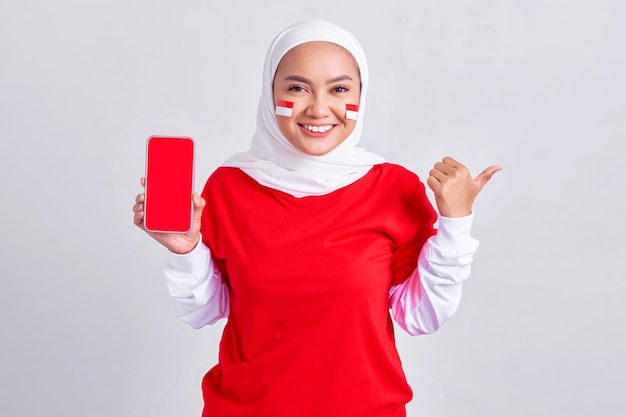 Allegra giovane donna musulmana asiatica in maglietta bianca rossa che mostra il telefono cellulare con schermo vuoto e indicando da parte isolato su sfondo bianco che celebra il giorno dell'indipendenza indonesiana il 17 agosto