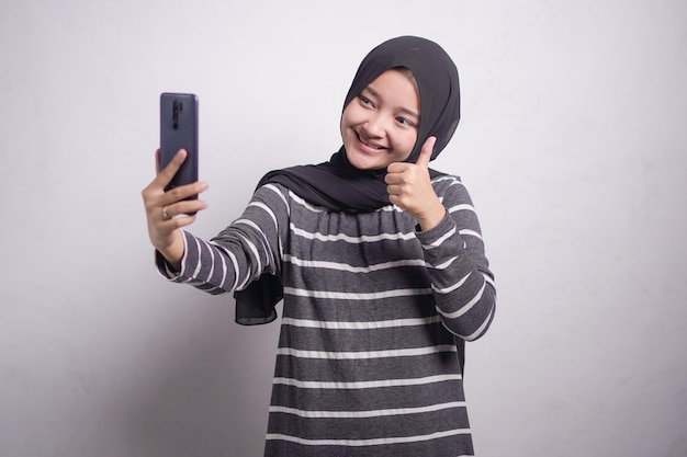 Allegra giovane donna musulmana asiatica in maglione bianco e nero facendo selfie girato sul telefono cellulare fare segno di pace isolato su sfondo bianco Concetto di stile di vita religioso della gente