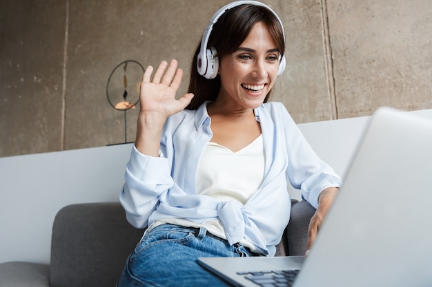 allegra giovane donna sorridente al chiuso a casa utilizzando il computer portatile con le cuffie sedersi sul divano in soggiorno salutando gli amici che parlano.