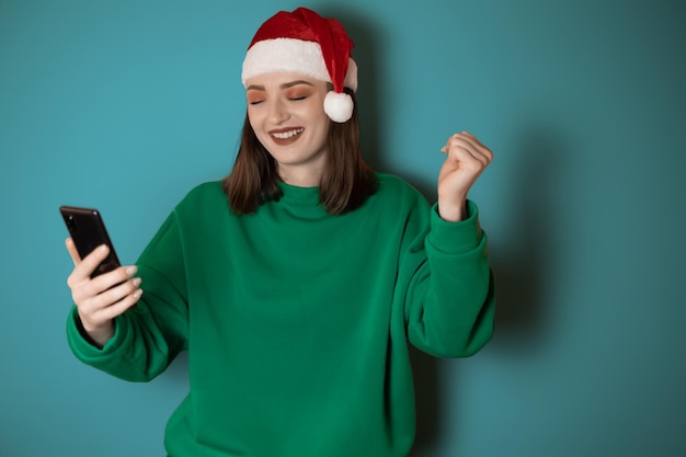 Allegra giovane donna vestita con un maglione verde e un cappello da Babbo Natale che tiene in mano un telefono cellulare facendo un gesto vincente isolato su uno sfondo verde Felice anno nuovo 2023 concetto di vacanza