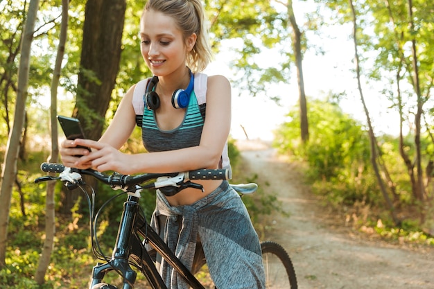 Allegra giovane ragazza fitness che trasporta zaino, indossa abbigliamento sportivo camminando con una bicicletta al parco, usando il telefono cellulare
