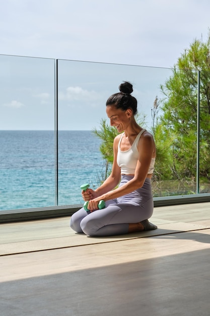 Allegra giovane sportiva in abbigliamento sportivo seduta con manubri in mano sul tappetino durante l'allenamento sulla terrazza contro il mare blu in sulight