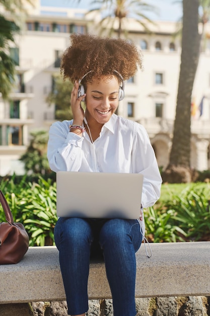 Allegra libera professionista donna nera che sorride e guarda lo schermo mentre lavora in remoto sul laptop e ascolta musica nel parco cittadino