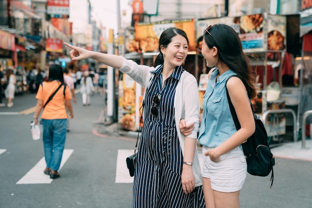 allegra ragazza asiatica sorride alla sua amica mentre indica in lontananza sulla strada con stand di snack nell'area urbana dello shopping durante l'estate.