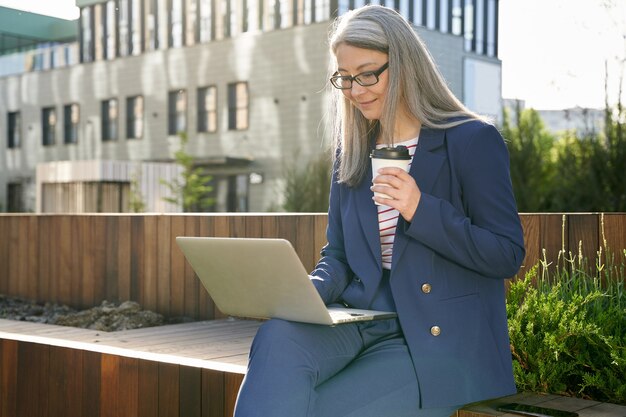 Allegra signora dai capelli grigi in abito da lavoro che tiene in mano una tazza di caffè mentre guarda lo schermo del laptop