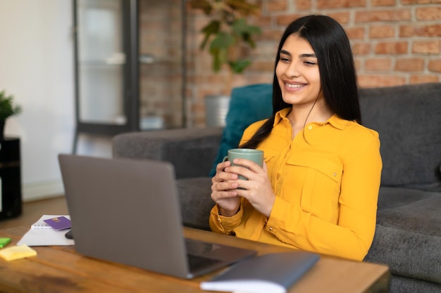 Allegra signora spagnola che guarda video sul laptop e beve caffè a casa guardando il computer