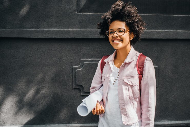 Allegra studentessa afro sorridente che indossa occhiali da vista si prepara per andare al college per la prima volta Felice giovane donna africana sorridente con zaino e rotolo di carta all'aperto Educazione della gente