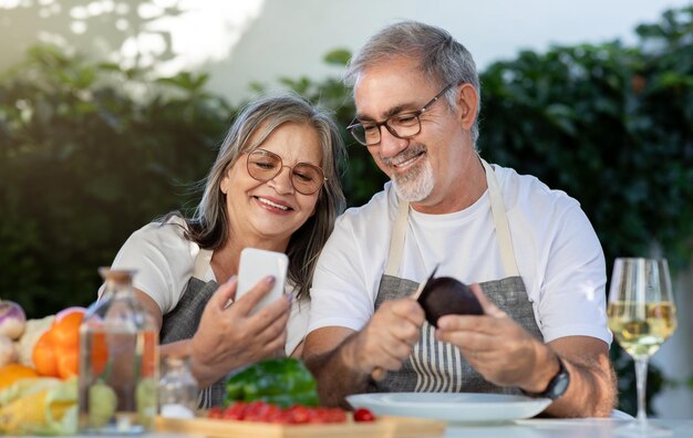 Allegra vecchia coppia europea in grembiuli con bicchieri di vino usa gli smartphone