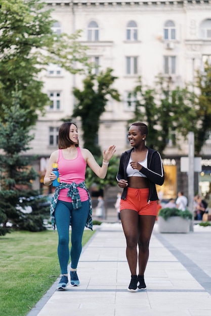 Allegri amici sorridenti in abbigliamento sportivo che camminano dopo una sessione di sport in città discutendo di donne multietniche che fanno jogging durante un allenamento fitness