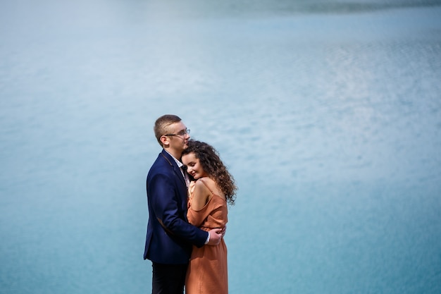 Allegri sposini vanno tenendosi per mano e ridendo, sullo sfondo di un lago e di un prato verde. Lo sposo allegro e la bella sposa con capelli ricci camminano nel prato