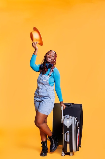 Allegro adulto in posa con cappello e bagaglio in studio, entusiasta dell'avventura delle vacanze con i trolley. Turista femminile che sorride alla macchina fotografica e si diverte durante il viaggio di vacanza.