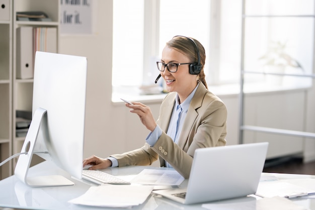 Allegro amichevole giovane rappresentante femminile call center in cuffia con microfono seduto al tavolo e utilizzando il computer durante la risoluzione del problema tecnico dei clienti