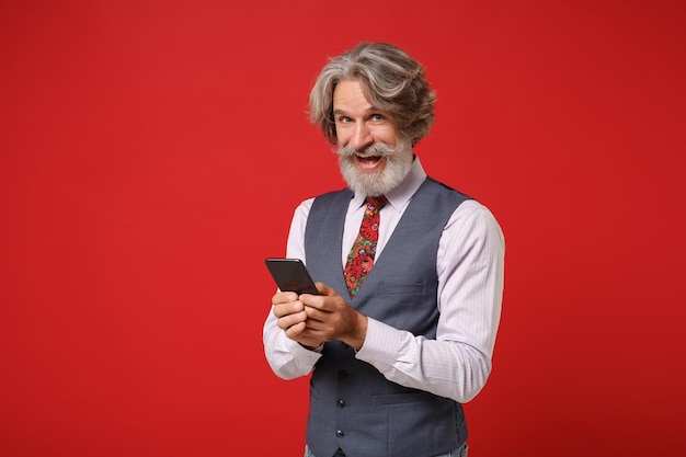 Allegro anziano uomo barbuto con baffi dai capelli grigi in camicia classica cravatta colorata in posa isolata su sfondo rosso in studio. Concetto di stile di vita delle persone. Mock up spazio di copia. Tenendo il cellulare.