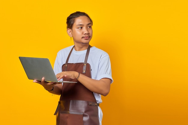 Allegro bel giovane barista che tiene in mano un laptop e guarda di traverso su sfondo giallo