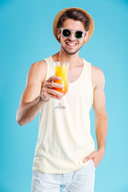 Allegro bel giovane con cappello e occhiali da sole con in mano un bicchiere di succo fresco sul muro blu