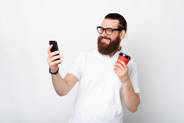 Allegro bell'uomo barbuto utilizza lo smartphone e tenendo la tazza di caffè su sfondo bianco