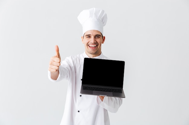 Allegro chef cuoco indossa uniforme in piedi sopra il muro bianco, tenendo il computer portatile dello schermo della banca