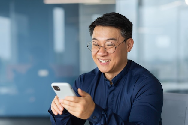 Allegro e sorridente uomo d'affari asiatico all'interno dell'ufficio utilizzando il messaggio di battitura a macchina dell'uomo del telefono e la navigazione