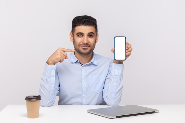 Allegro elegante giovane uomo d'affari seduto nel posto di lavoro dell'ufficio che punta al telefono cellulare con schermo vuoto mock up display vuoto per la pubblicità studio indoor girato isolato su sfondo bianco