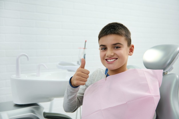 Allegro fiducioso coraggioso bel bambino di età elementare, ragazzo di scuola mostra il pollice in su e sorride con un bel sorriso a trentadue denti guardando la fotocamera mentre è seduto sulla poltrona del dentista dopo il trattamento dentale