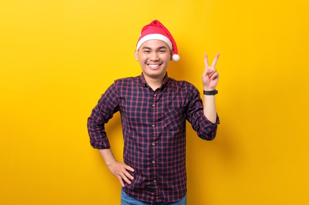 Allegro giovane asiatico con cappello da Babbo Natale che mostra il segno della vittoria guardando la fotocamera su sfondo giallo dello studio Happy New Year 2023 celebrazione buon concetto di vacanza