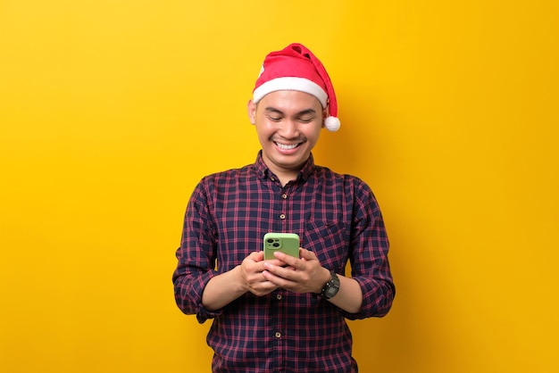 Allegro giovane asiatico con cappello da Babbo Natale che utilizza lo smartphone per inviare messaggi con gli amici su sfondo giallo studio Happy New Year 2023 celebrazione buon concetto di vacanza
