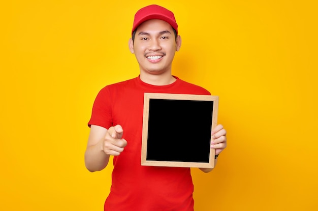 Allegro giovane asiatico in berretto rosso tshirt uniforme dipendente lavora come rivenditore corriere in possesso di lavagna vuota che punta il dito alla fotocamera su sfondo giallo Concetto di servizio di consegna professionale