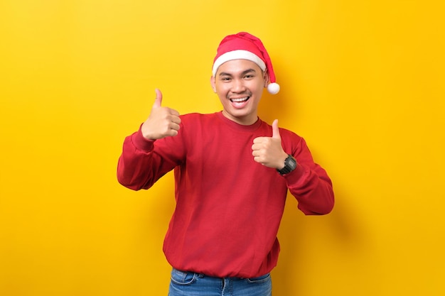 Allegro giovane asiatico in cappello di Babbo Natale che mostra i pollici in su guardando la fotocamera su sfondo giallo studio celebrazione vacanze di Natale e concetto di Capodanno