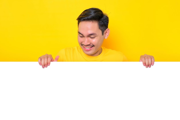 Allegro giovane asiatico in maglietta casual che guarda il tabellone pubblicitario bianco isolato su sfondo giallo Concetto di cartellone promozionale