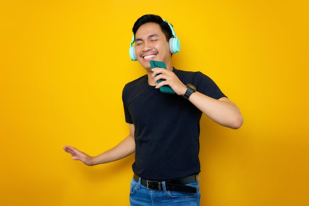Allegro giovane asiatico in maglietta casual in cuffia cantando la sua canzone preferita utilizzando il telefono cellulare come microfono isolato su sfondo giallo Concetto di stile di vita di emozioni della gente