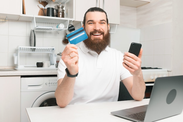 Allegro giovane barbuto hipster uomo seduto in cucina e pagare online con carta di credito e smartphone