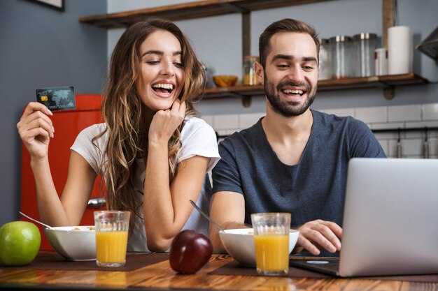 Allegro giovane coppia shopping con laptop e carta di credito mentre si consuma la colazione