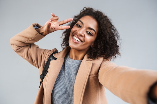 Allegro giovane donna africana che indossa cappotto in piedi isolato sopra il muro grigio, prendendo un selfie, gesto di pace