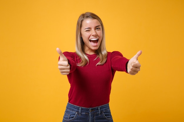 Allegro giovane donna bionda ragazza in abbigliamento casual in posa isolato sul ritratto in studio di sfondo muro giallo arancio. Concetto di stile di vita di emozioni della gente. Simula lo spazio della copia. Mostrando i pollici in su, sbattendo le palpebre.