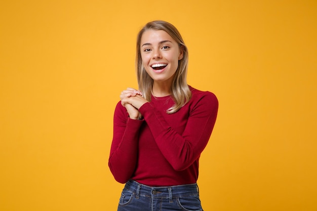 Allegro giovane donna bionda ragazza in abiti casual in posa isolato su sfondo giallo arancione ritratto in studio. Concetto di stile di vita di emozioni sincere della gente. Simula lo spazio della copia. Tenendo le mani giunte.