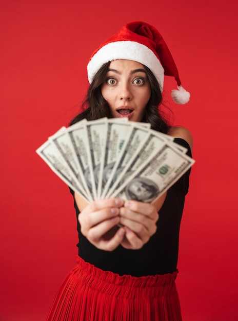 Allegro giovane donna che indossa il cappello di Natale in piedi isolato su rosso, mostrando le banconote in denaro