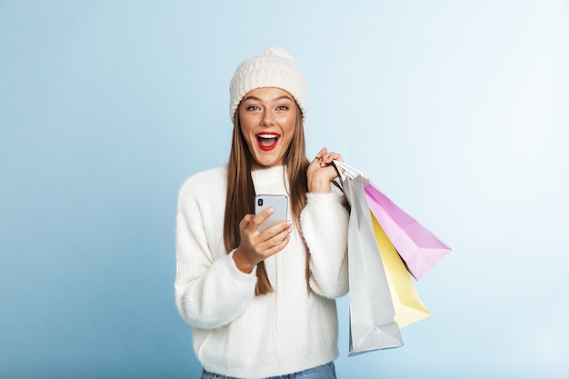 Allegro giovane donna che indossa un maglione, tenendo il telefono cellulare, portando le borse della spesa