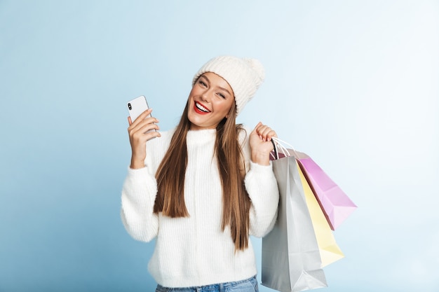 Allegro giovane donna che indossa un maglione, tenendo il telefono cellulare, portando le borse della spesa