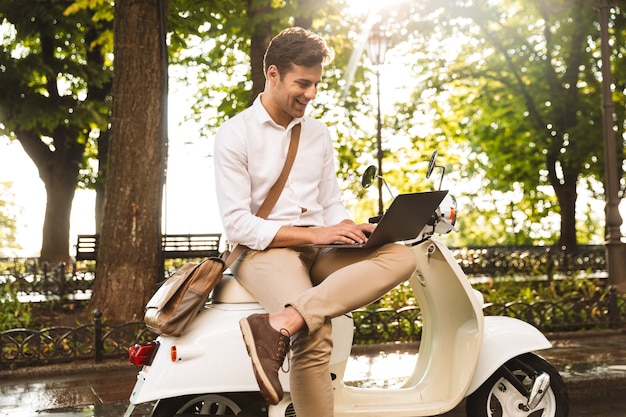 Allegro giovane imprenditore seduto su una moto all'aperto, lavorando su un computer portatile