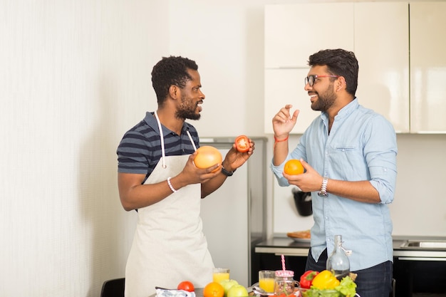 Allegro giovane indiano con gli occhiali e uomo afroamericano che indossa un grembiule che si diverte in cucina. Due amici maschi in piedi vicino alla scrivania con frutta e verdura e ridono mentre cucinano la colazione.