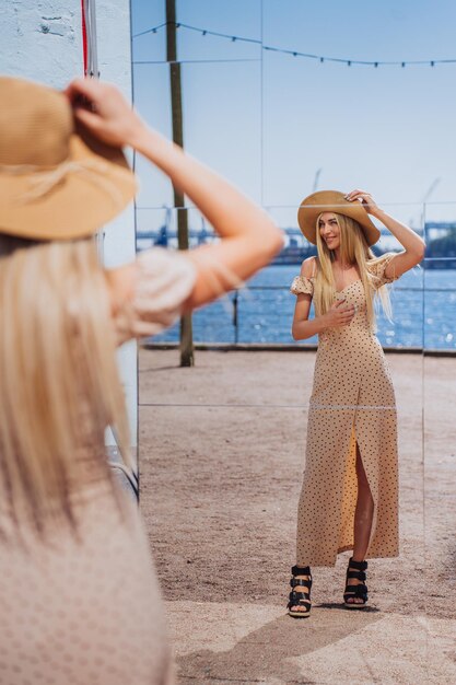 Allegro giovane modello femminile svedese in piedi fuori cappello adatto guardando allo specchio godendo le vacanze estive Donna caucasica in viaggio in abito beige che cammina in riva al mare Persone di successo