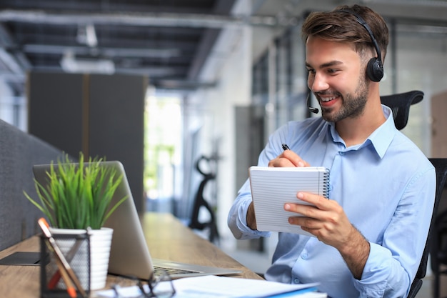 Allegro giovane operatore telefonico maschio di supporto in cuffia, sul posto di lavoro durante l'utilizzo di laptop, servizio di assistenza e concetto di call center di consulenza clienti.