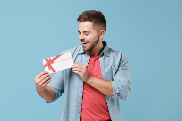 Allegro giovane ragazzo barbuto 20s in camicia casual in posa isolato su sfondo blu pastello parete ritratto in studio. Persone sincere emozioni concetto di stile di vita. Mock up spazio di copia. Tieni il buono regalo.