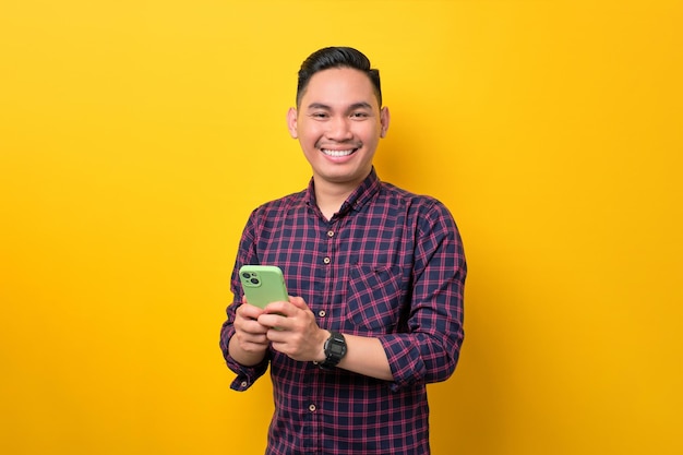 Allegro giovane uomo asiatico che tiene smartphone e sorride alla macchina fotografica isolata su sfondo giallo