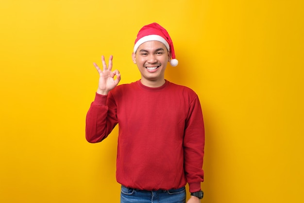 Allegro giovane uomo asiatico in cappello di Babbo Natale che mostra segno ok e guardando la fotocamera su sfondo giallo studio celebrazione vacanze di Natale e concetto di Capodanno