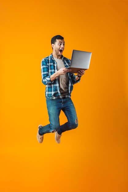 Allegro giovane uomo che indossa la camicia a quadri saltando isolato su sfondo arancione, utilizzando il computer portatile