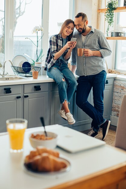 Allegro giovane uomo e donna che sorridono in cucina e guardano lo schermo di uno smartphone