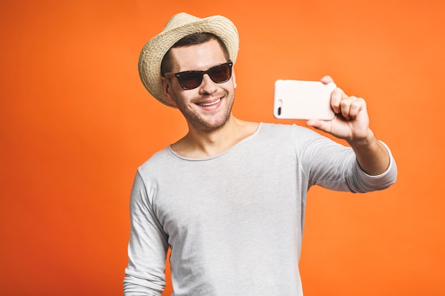 Allegro giovane uomo in cappello e occhiali da sole tenendo selfie con lo smartphone isolate su sfondo arancione