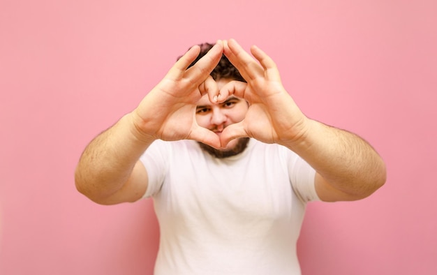 Allegro giovane uomo in sovrappeso che mostra il gesto del cuore alla fotocamera su sfondo rosa e sorridente